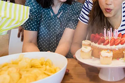 Bild från ett födelsedagskalas där en kvinna blåser ut ljus från en tårta i bakgrunden, och en stor skål med chips i förgrunden.