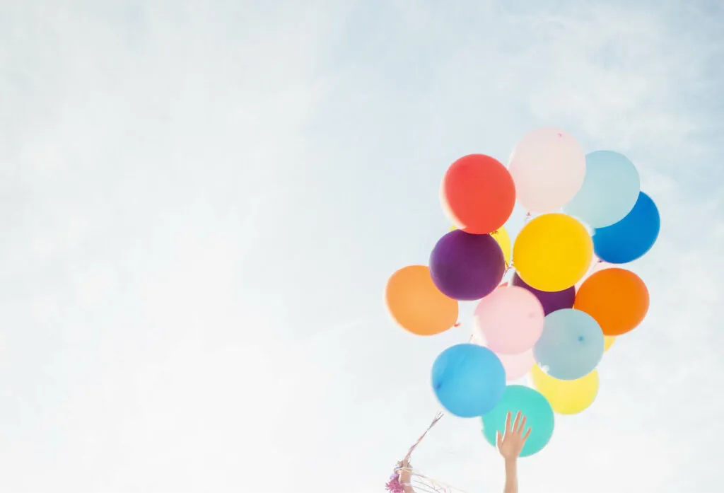 Händer som håller en stor samling flygande ballonger i olika färgglada färger mot en ljusblå himmel.