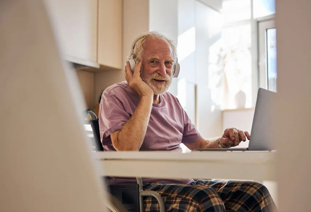 En äldre man i rullstol sitter vid sitt köksbord och lyssnar på musik, och tittar glatt in i kameran. Symbolisk bild för Sociala Missionens seminarier på temat hälsa och livsmod hos äldre. Det här seminariet med fokus på musikens betydelse för hälsan och att främja livsmod hos äldre.