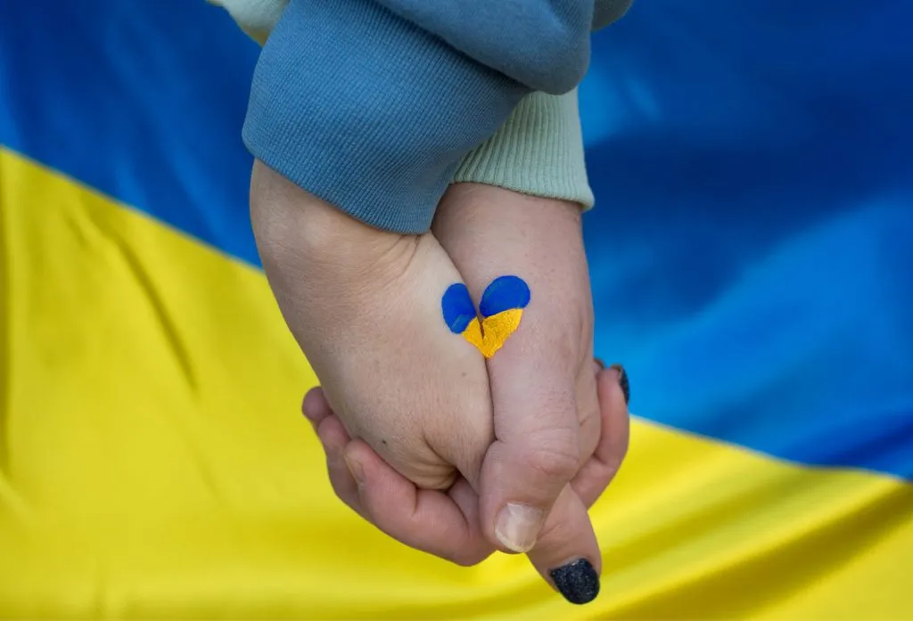 Ukrainska flaggan i bakgrunden, och i förgrunden två målade händer (en vuxen och ett barn) som håller i varandra som tillsammans bildar ett hjärta med Ukrainska färgerna gult och blått.