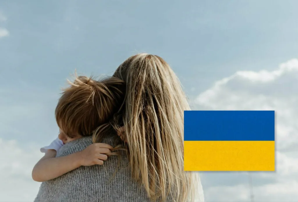 En mamma som kramar om sitt barn som hänger över mammans axel. Över bilden på mamman och barnet finns i högra hörnet i nedre kant en ukrainsk flagga. Symbolisk bild för Webbinariet om Ukraina och massflyktsdirektivet.