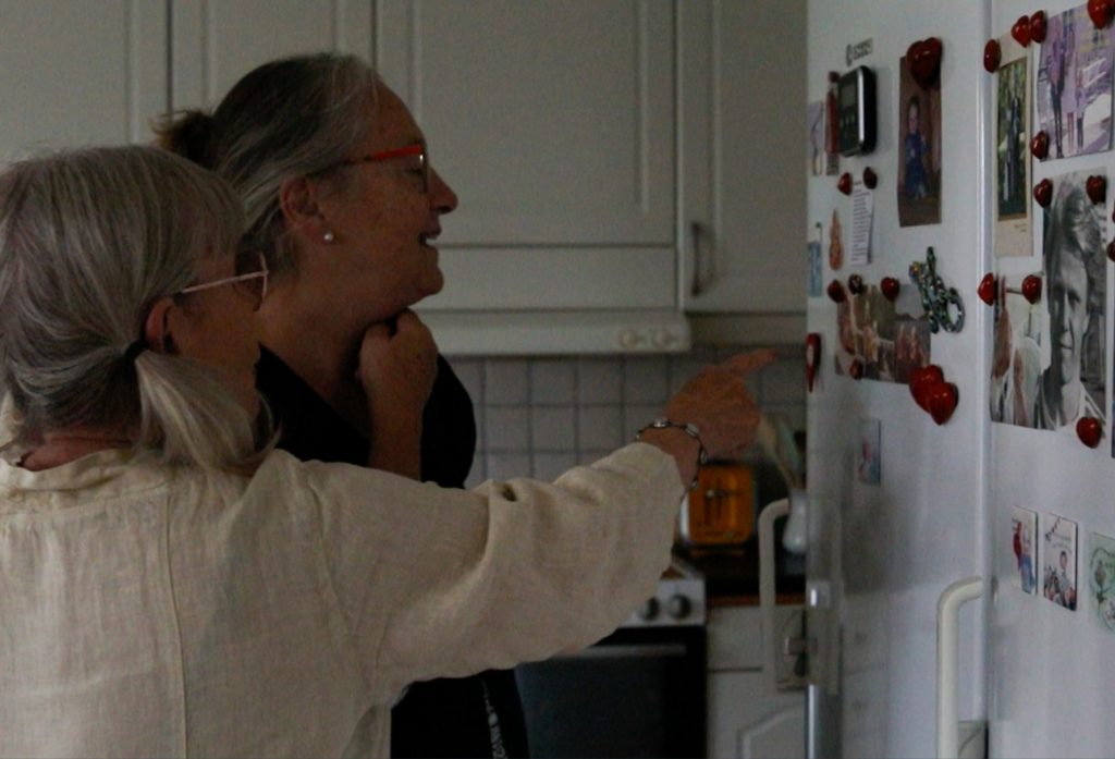 Katarina Grape (tidigare deltagare i en samtalsgrupp för äldre på Gotland) i förgrunden, pekar mot sitt kylskåp och visar Lilian (gruppledare för samtalsgruppen Katarina deltog i) bilder från hennes liv som sitter på kylskåpet.