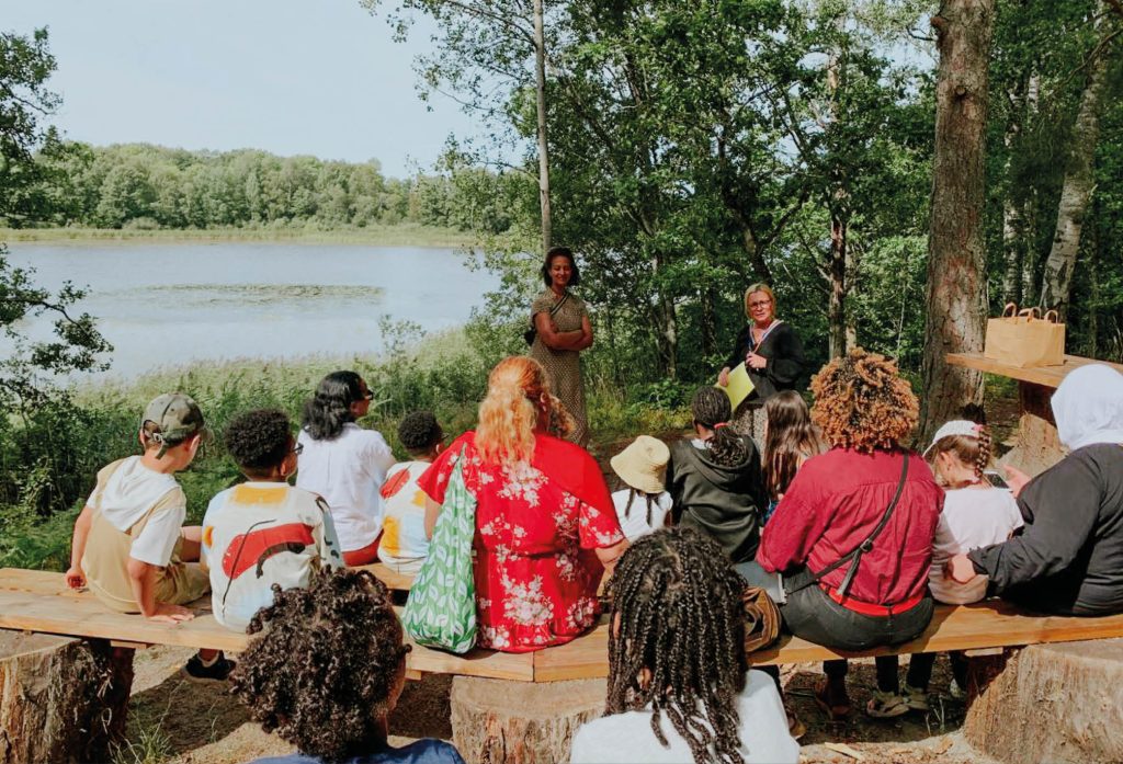 Bild från Sociala Missionens sommarläger för barn och familjer som som lever i utsatthet i Stockholm. På bilden syns vatten i bakgrunden, och grönskande träd, med flera familjer och barn som sitter på bänkar och tittar mot gruppledarena Yodit och Lotta som presenterar en lek de ska göra tillsammans.