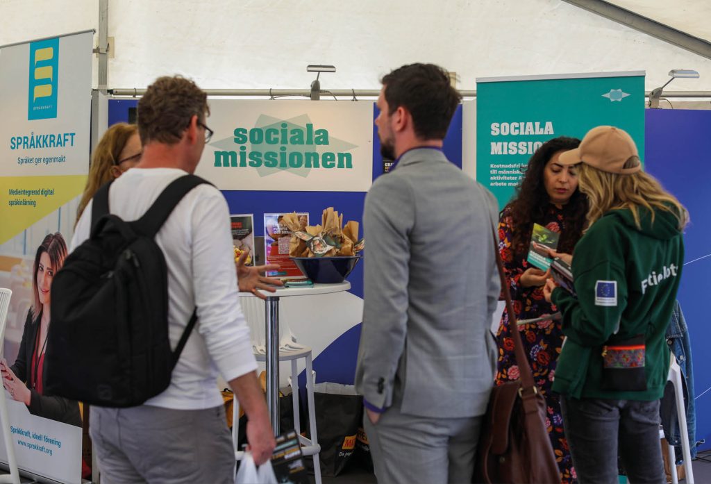 Bild från Sociala Missionens monterplats i tältet för Civilsamhället under Järvaveckan 2024, där Sociala Missionens personal står och svarar på frågor till nyfikna besökare. I monterplatsen syns, utöver besökande människor, Sociala Missionens logga och en stor skål med potatis och en med chips, och innehåll som berättar mer om Sociala Missionen och vår historia.