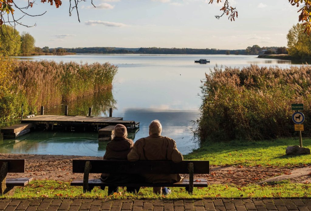 Bild bakifrån på en äldre man och äldre kvinna som sitter på en bänk och tittar ut över en vacker, stilla sjö. Runt omkring dem finns grönska/natur som håller på att övergå till höst. Bilden är omslagsbild för projektet "Livet här och nu" som startar hösten 2025 i Åbybergskyrkan och är en samtalsgrupp som riktar sig till människor med tidig demens / kognintiv sjukdom.