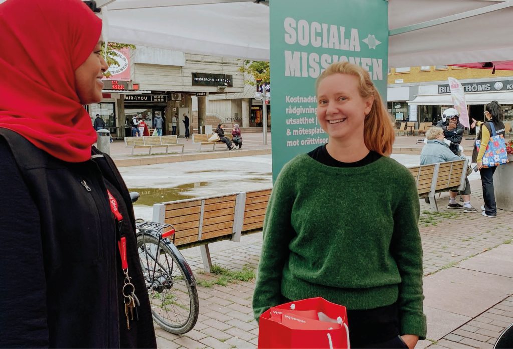 Bild på Karin, Sociala Missionens nya rådgivare för frågor som rör äldre personer i utsatthet, under seniordagarna på Skärholmen 2025 när hon står på Skärholmens torg under ett samtal med en kvinna från annan organisation samtidigt som hon tittar in i kameran och ler.